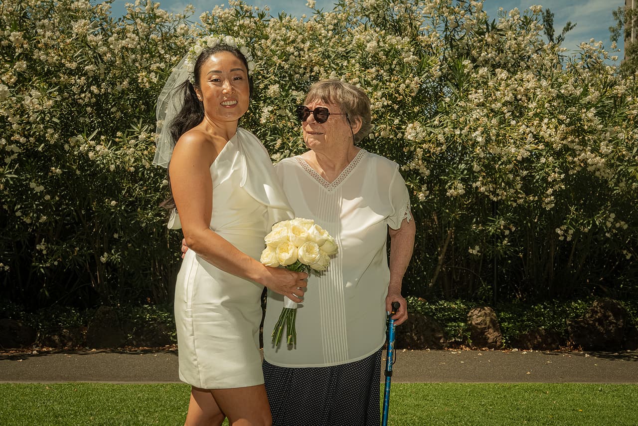 Cecile arm in arm with an older guest in sunglasses on the lawn in front of the flowering hedge