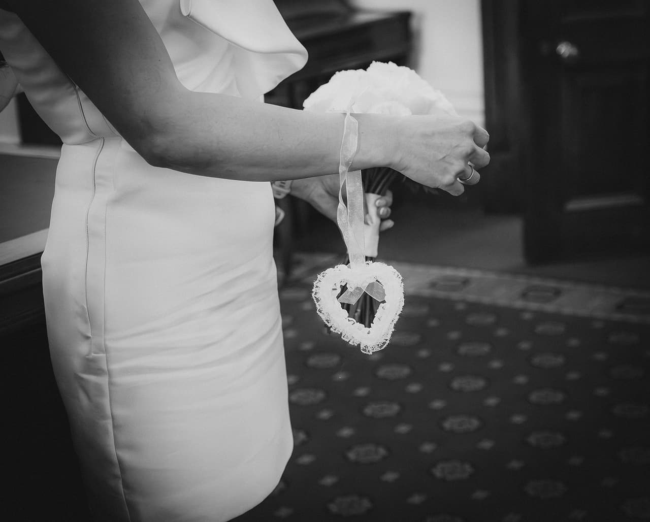 Black and white close up of a small white lace heart hanging from Cecile’s hand beside her dress