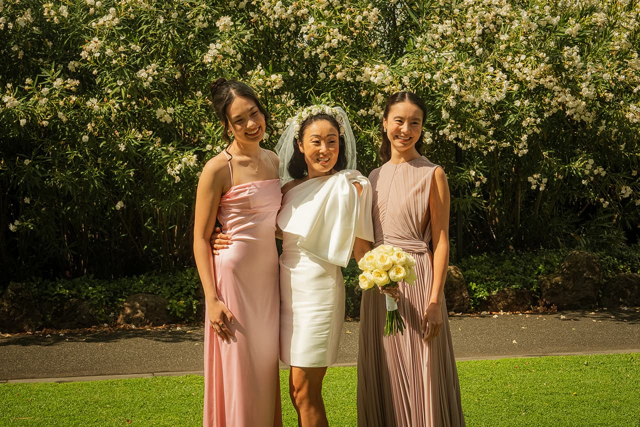 Cecile with two friends in long pink and mauve dresses in front of a flowering hedge