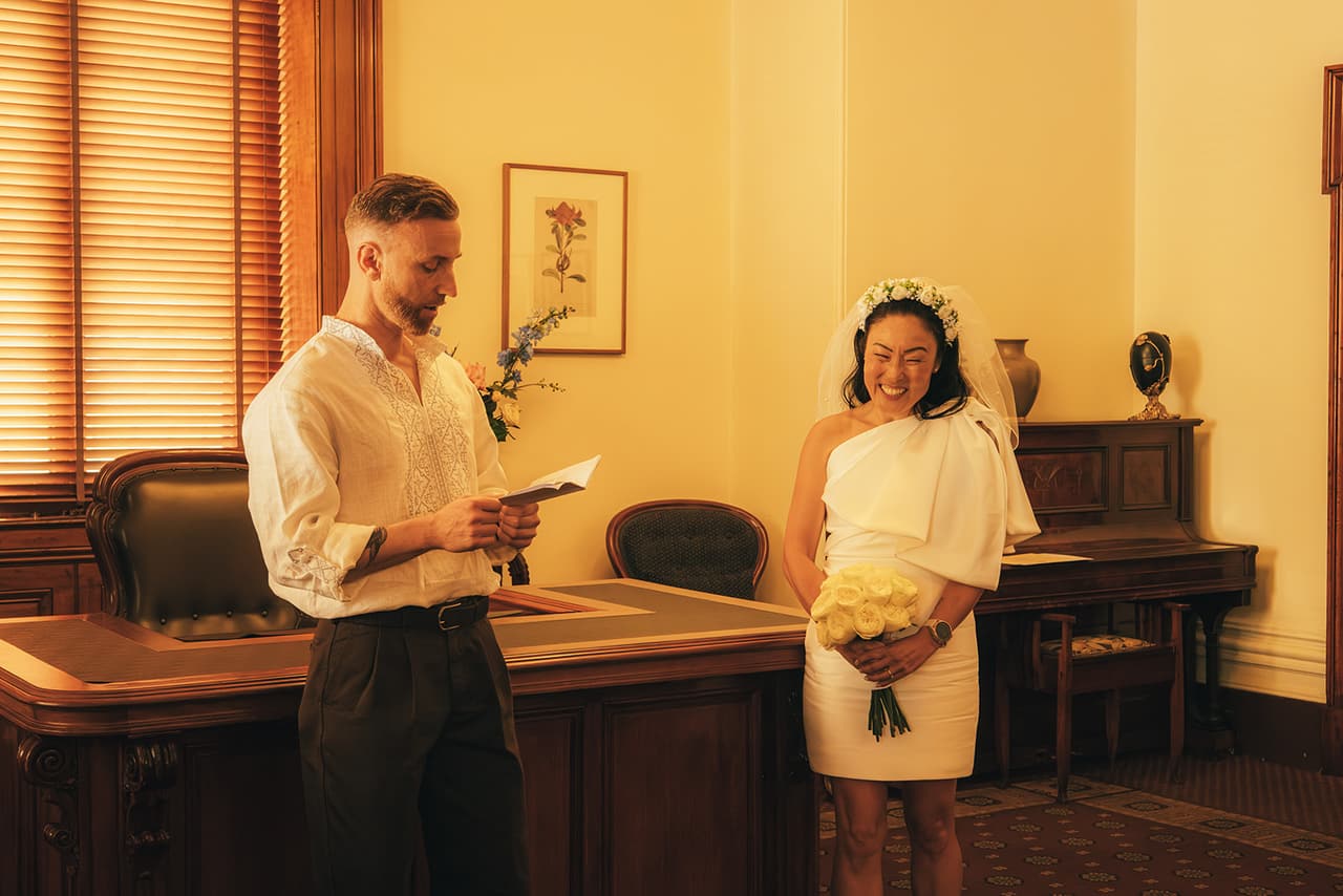 Stefan reading his vows from a card as Cecile laughs beside the registry desk