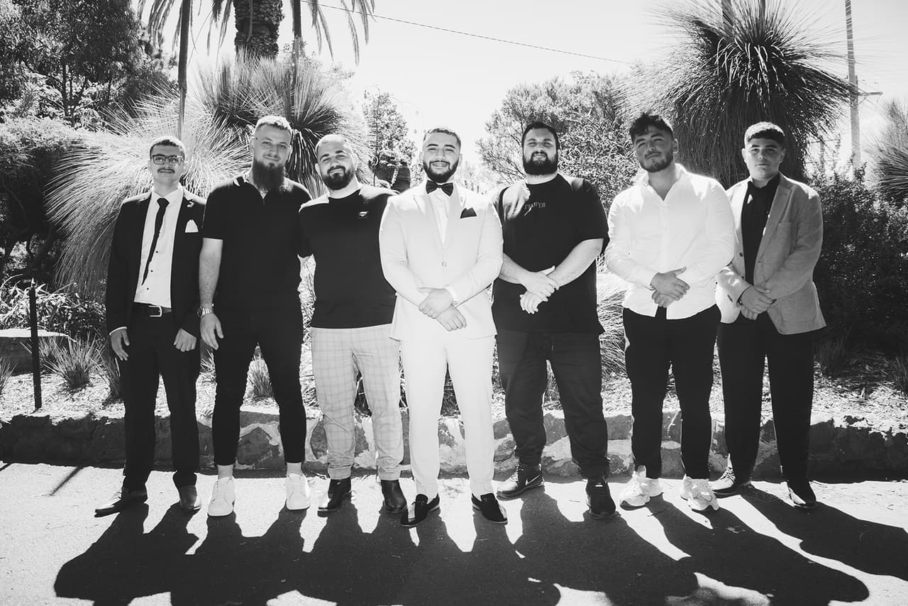 Black and white photo of Yasir standing in a row with six friends on a sunny garden path, their long shadows in front of them