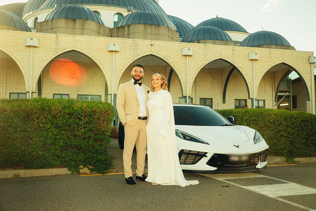 Elektra and Yasir in front of the white domes and arches of the Sunshine Mosque, a white sports car beside them