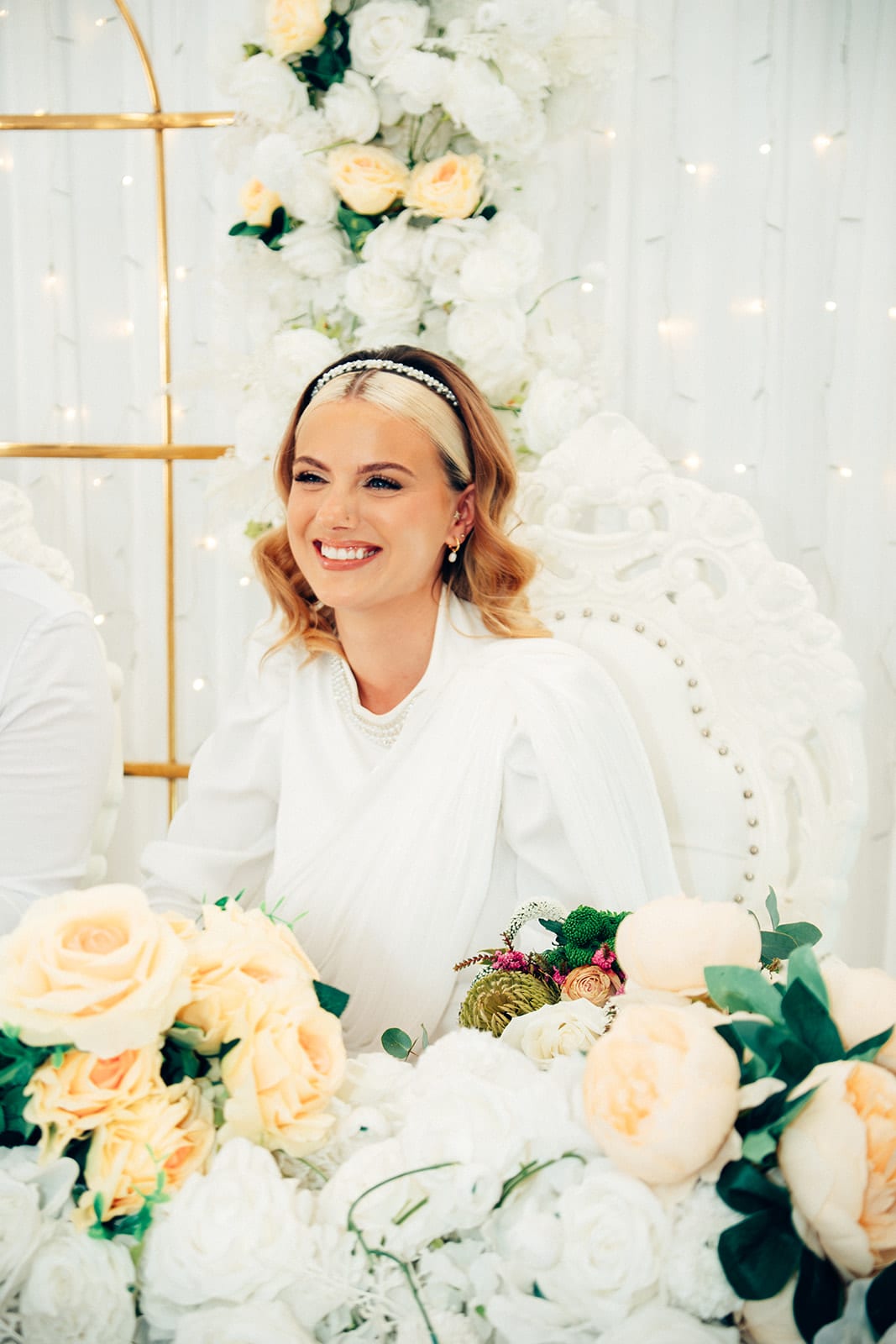 Elektra laughing at the head table behind white and peach roses, the gold arches behind her