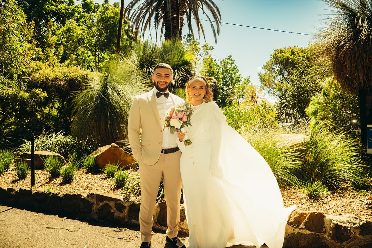 Elektra and Yasir standing among palms and grass trees in a sunny garden