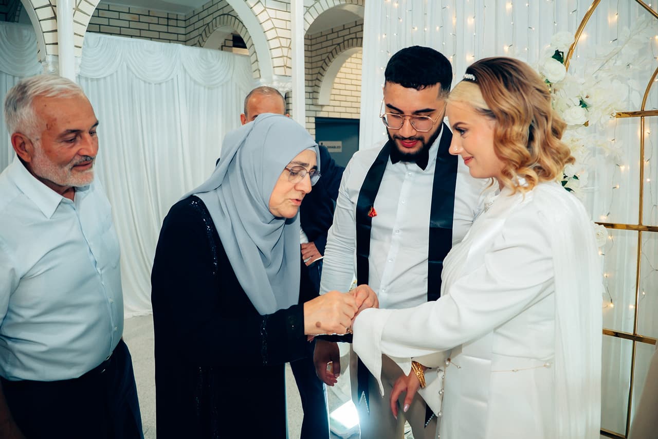 An older woman in a grey headscarf fastening a bracelet on Elektra’s wrist as an older man smiles beside them