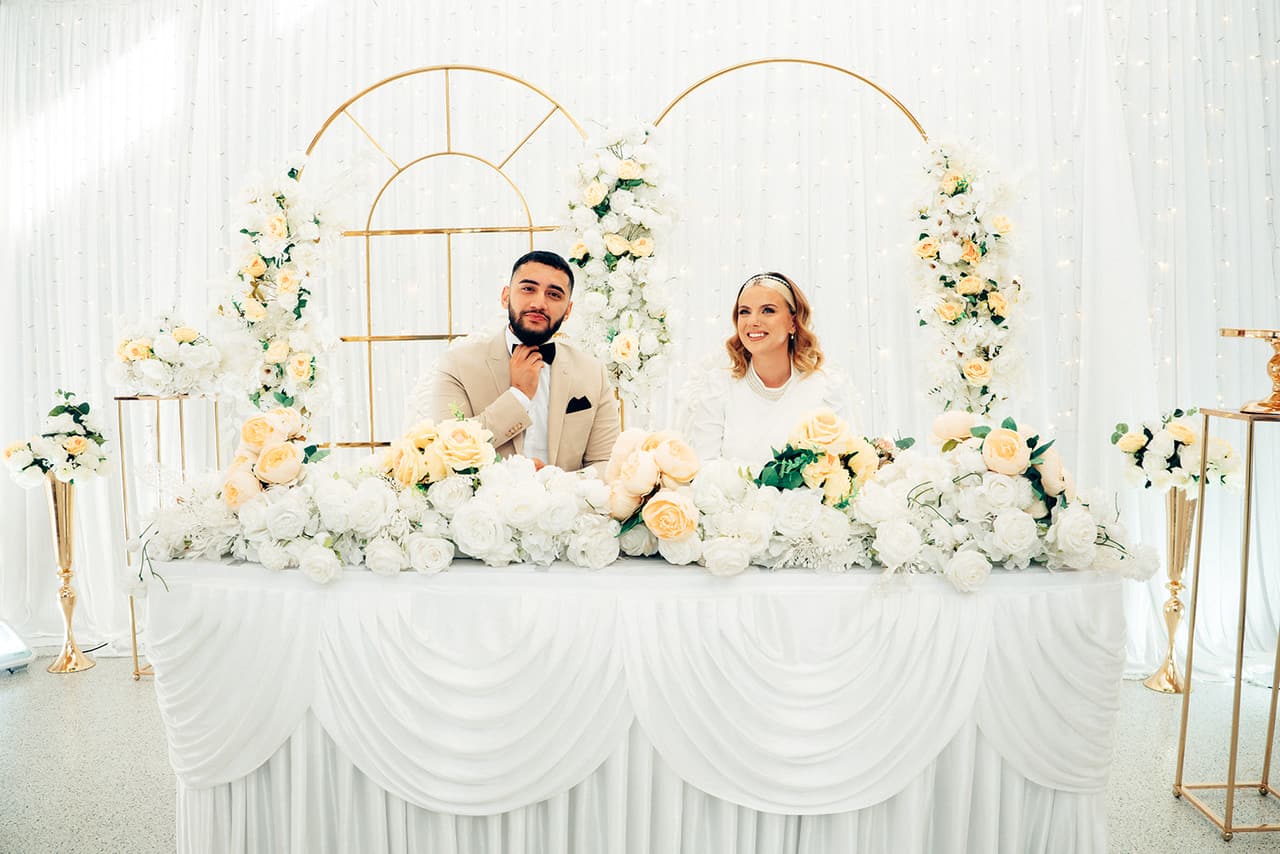 The head table under a gold arch and white flowers in the reception hall