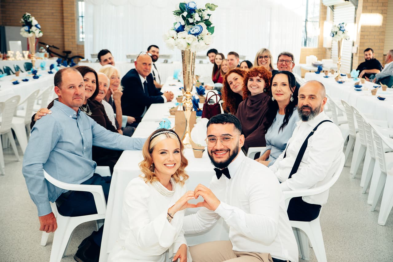 Elektra and Yasir making heart shapes with their hands at a long table of smiling guests