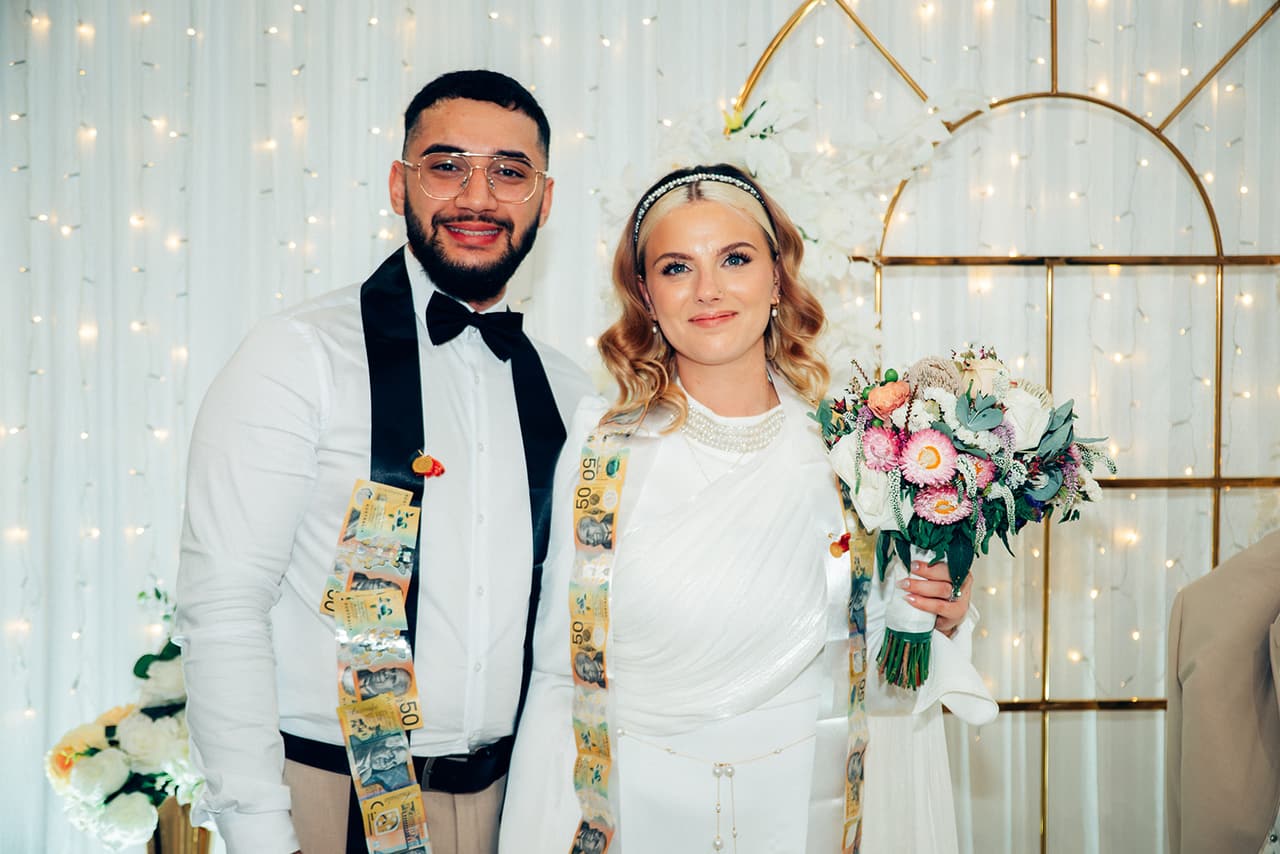 Yasir and Elektra smiling with sashes of pinned banknotes over their shoulders, her bouquet in her hand