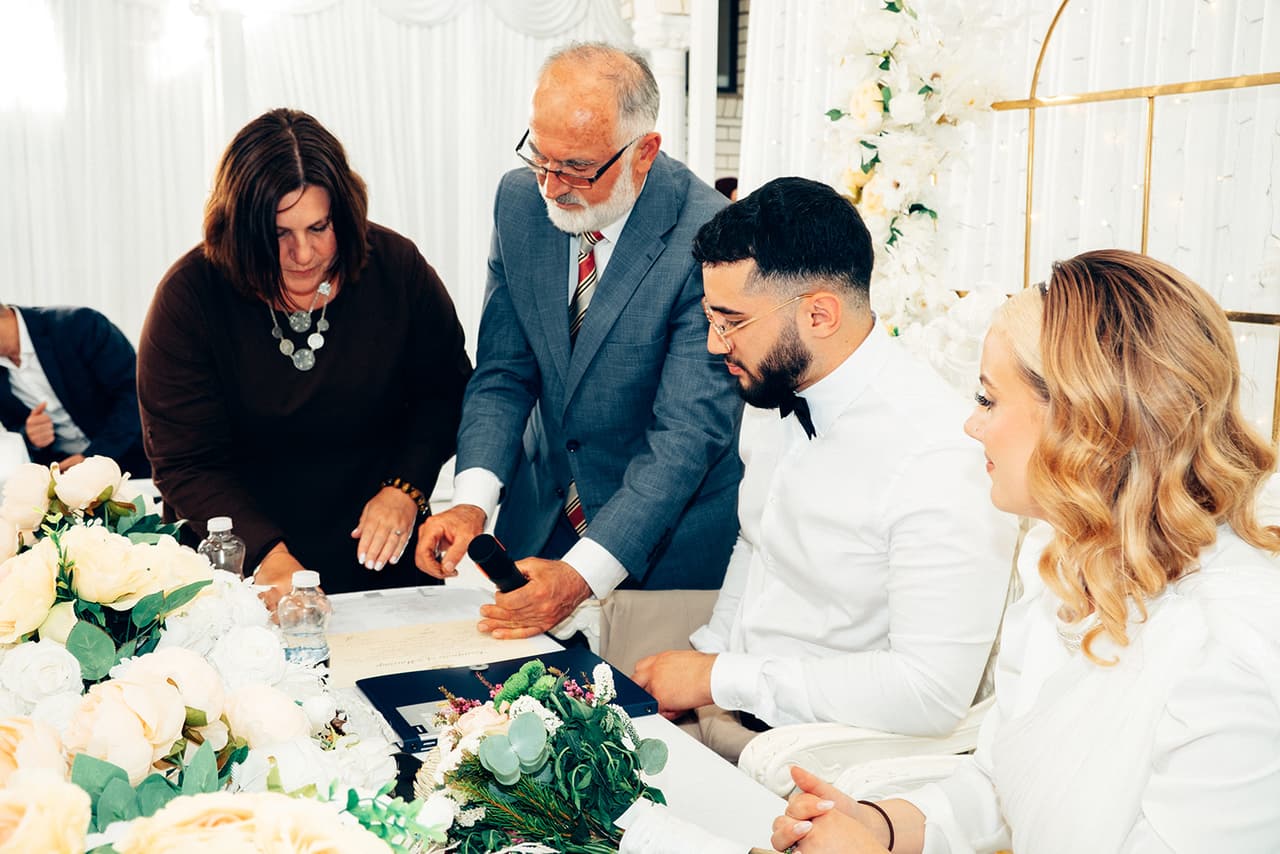 An older man in a grey suit and a woman in brown showing Yasir where to sign at the head table as Elektra watches