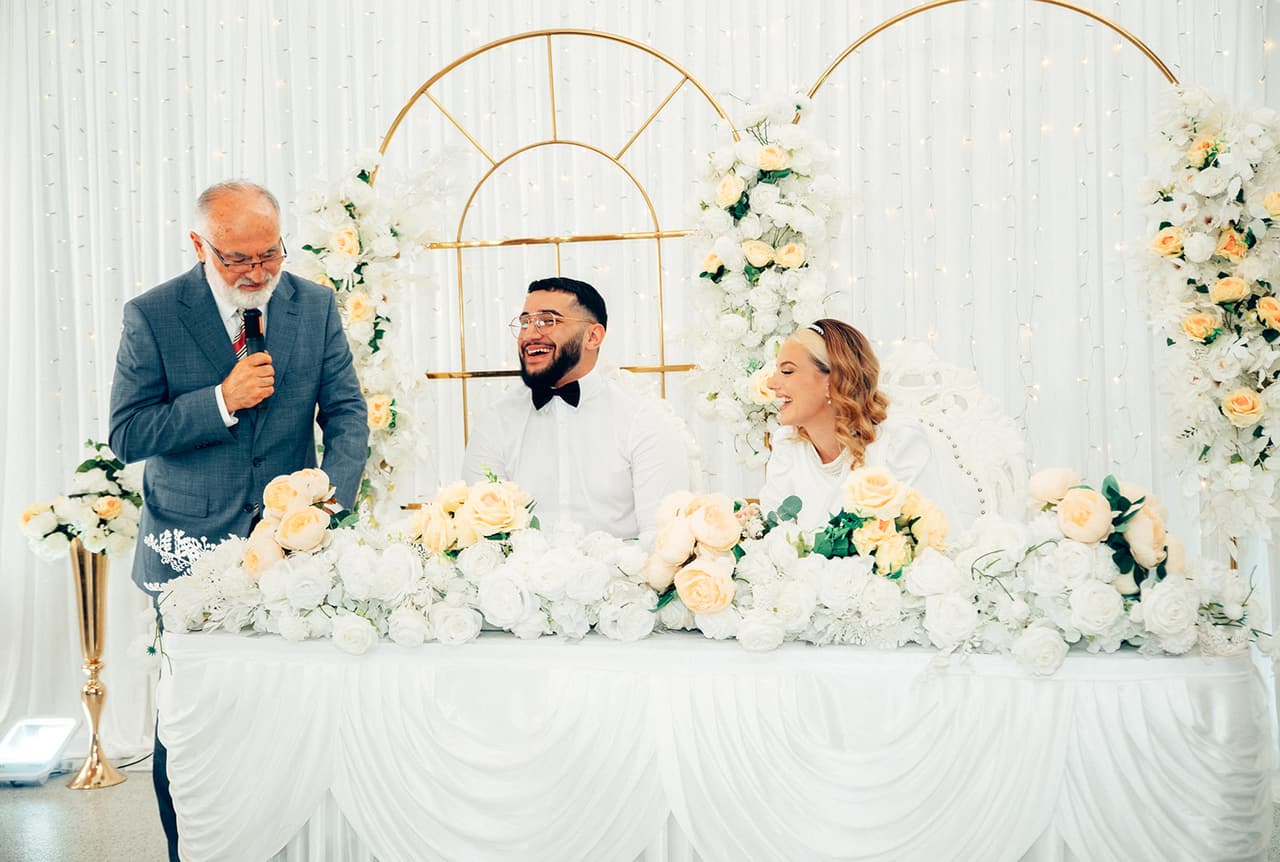 An older man in a grey suit speaking into a microphone in front of the head table as Yasir laughs and Elektra smiles