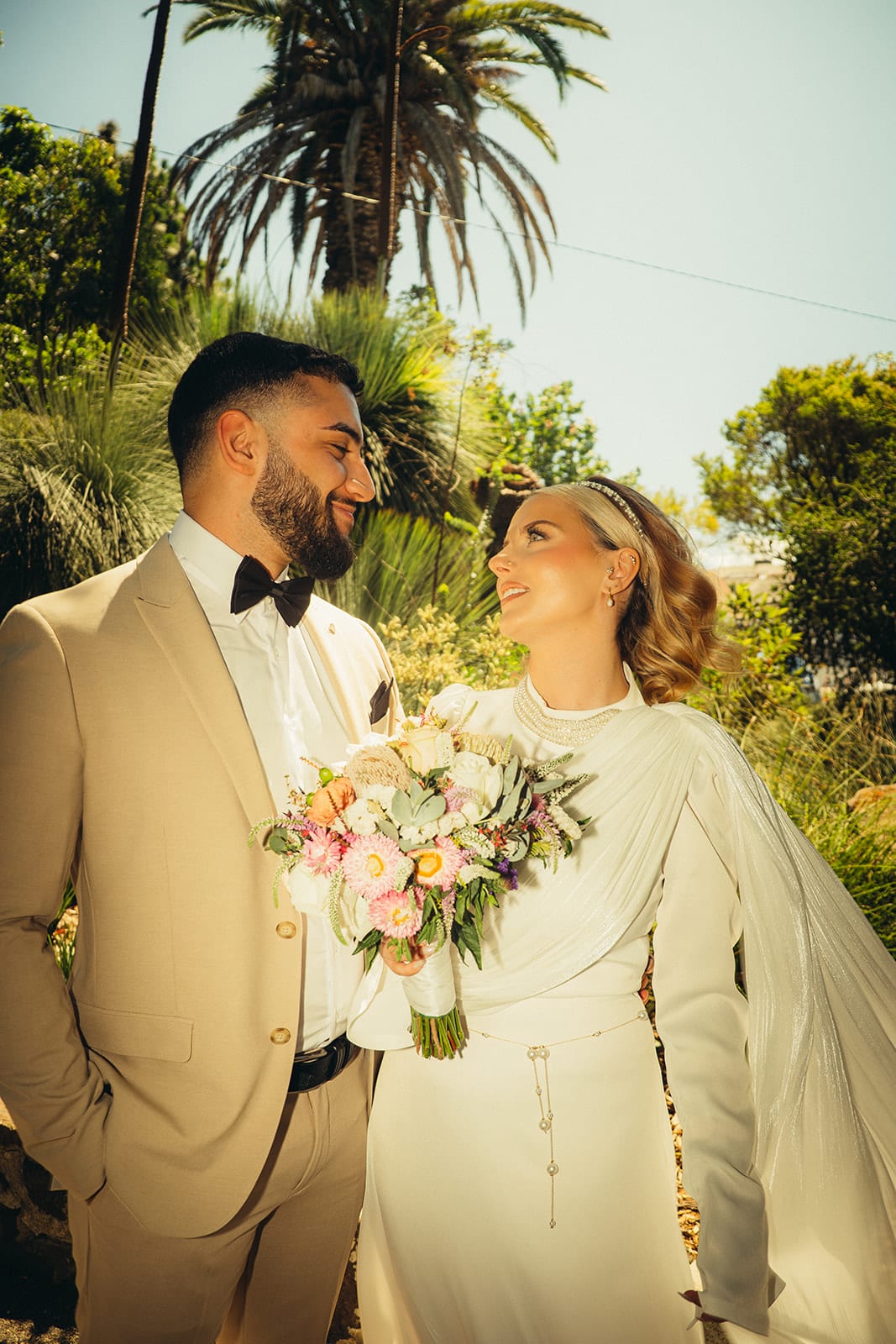 Yasir and Elektra smiling at each other among palms and grass trees, a bouquet of pink and peach roses between them