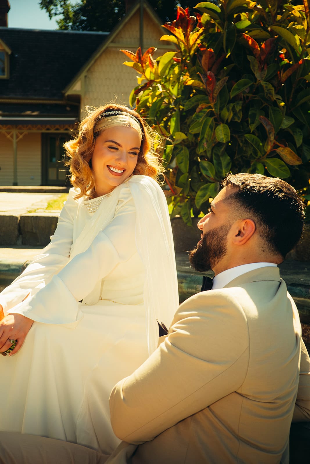 Elektra laughing as she and Yasir sit on the edge of a round stone pond, an old cottage with a decorated gable behind them