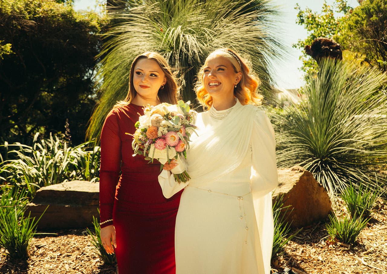 Elektra laughing beside a woman in a red dress among the grass trees