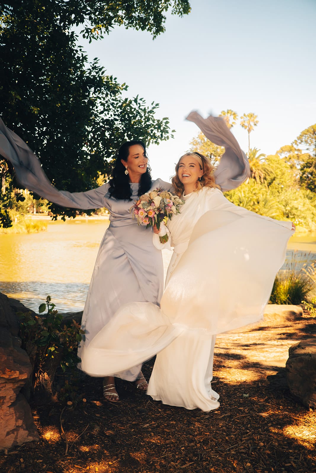 Elektra and a friend in a silver satin dress swirling their dresses and sleeves beside the lake