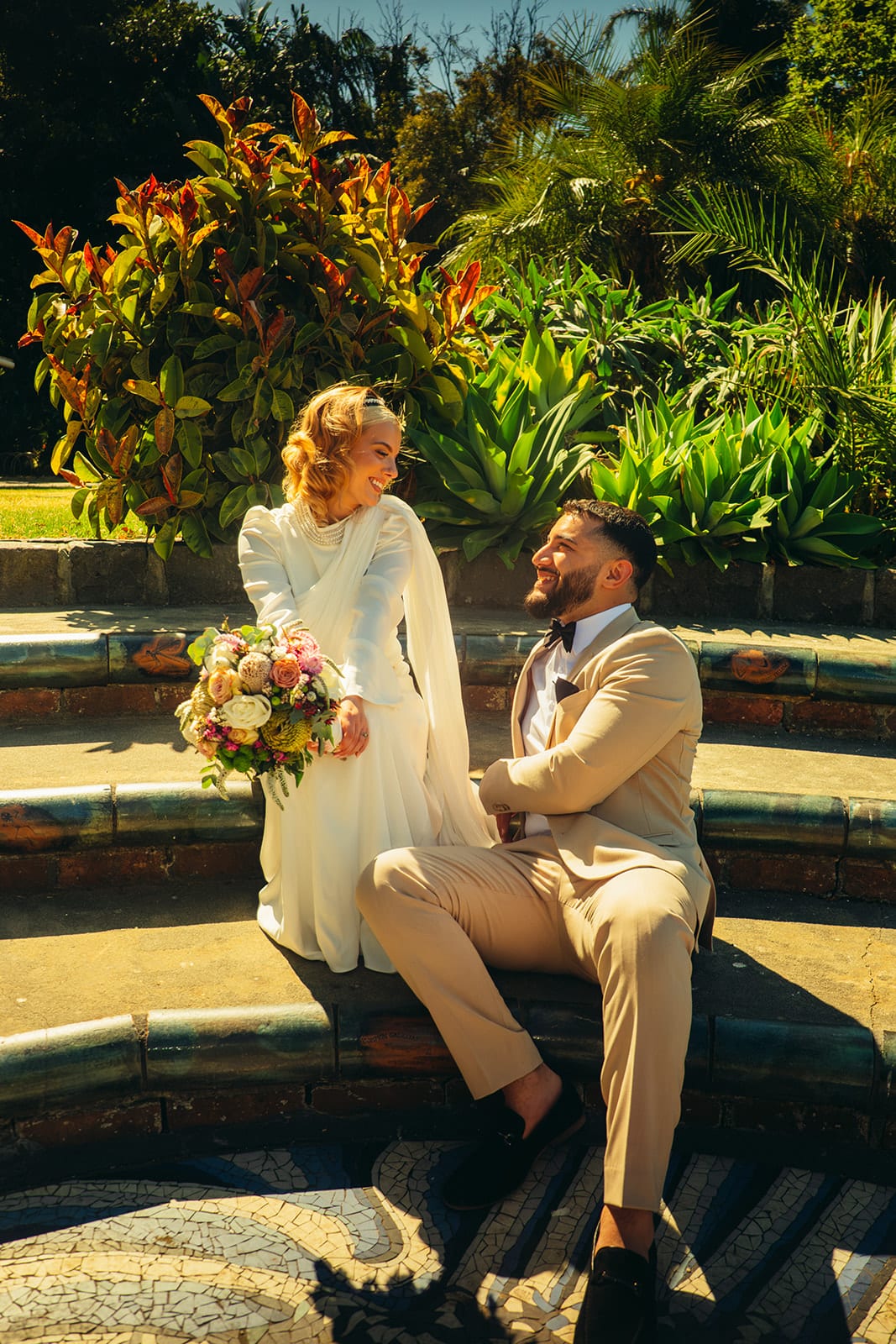 Yasir sitting on the edge of a garden fountain as Elektra stands beside him