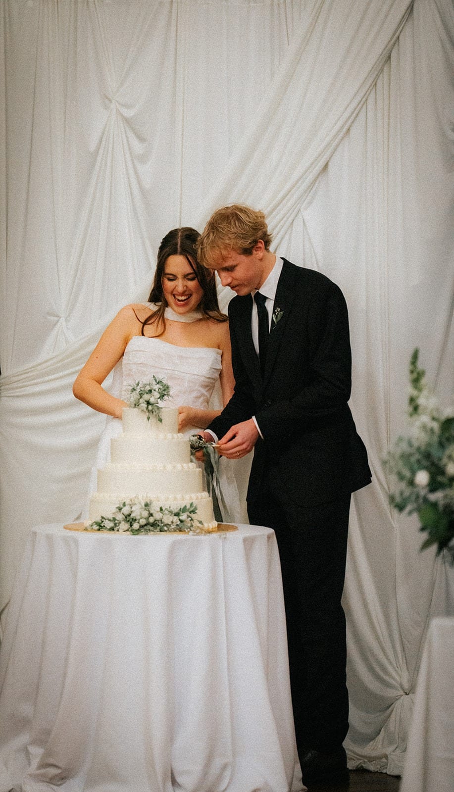 Eli and Charlotte cutting their cake in front of white draped fabric