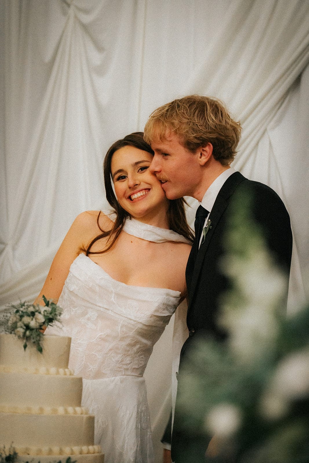 Charlotte smiling as Eli kisses her cheek beside the cake