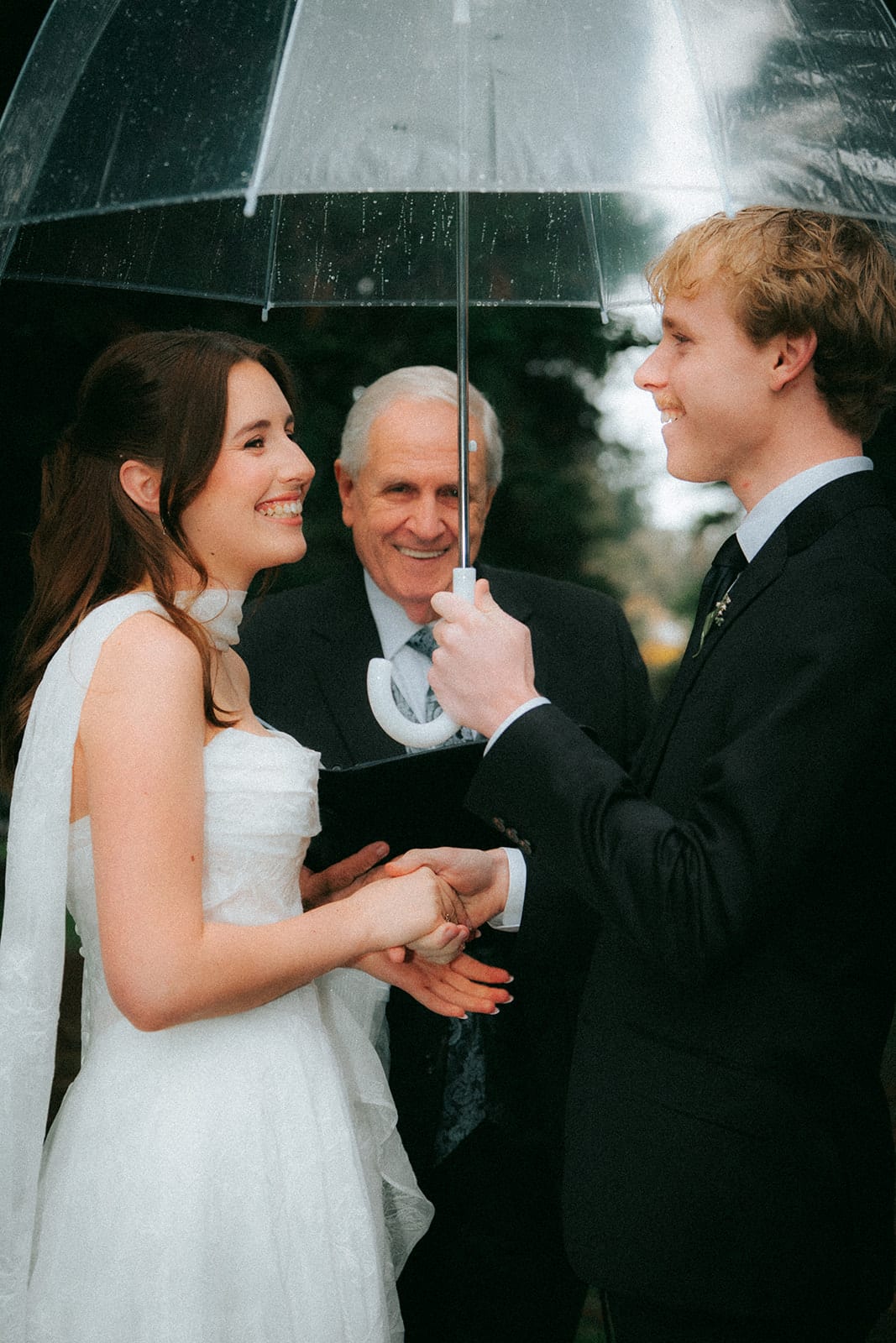 Charlotte and Eli smiling at each other under the clear umbrella the celebrant holds over them