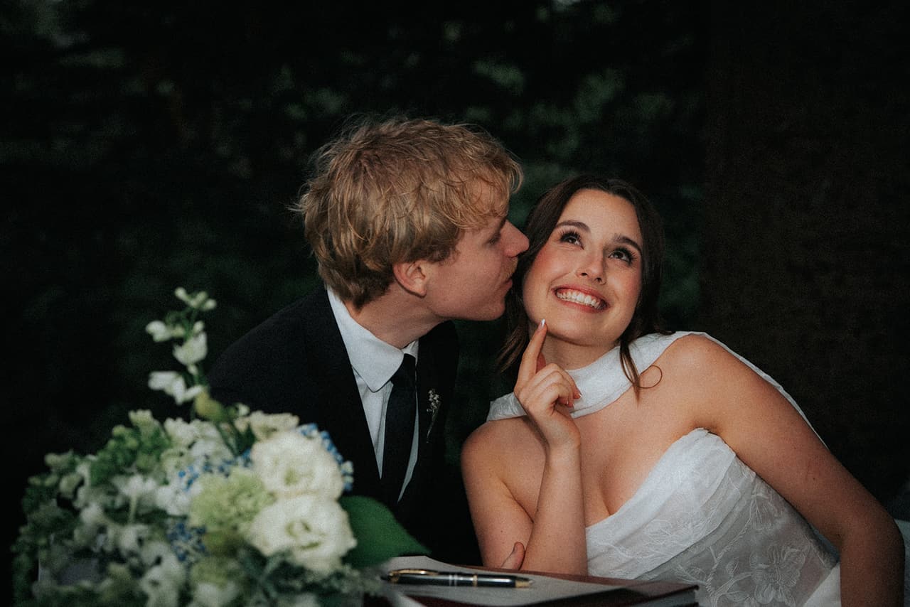Charlotte grinning upwards as Eli kisses her cheek at the signing table