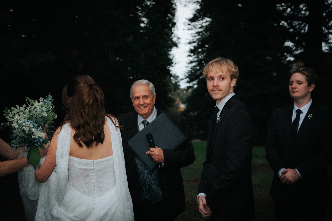 Eli turning to Charlotte as she reaches the front, the celebrant smiling beside him with his folder