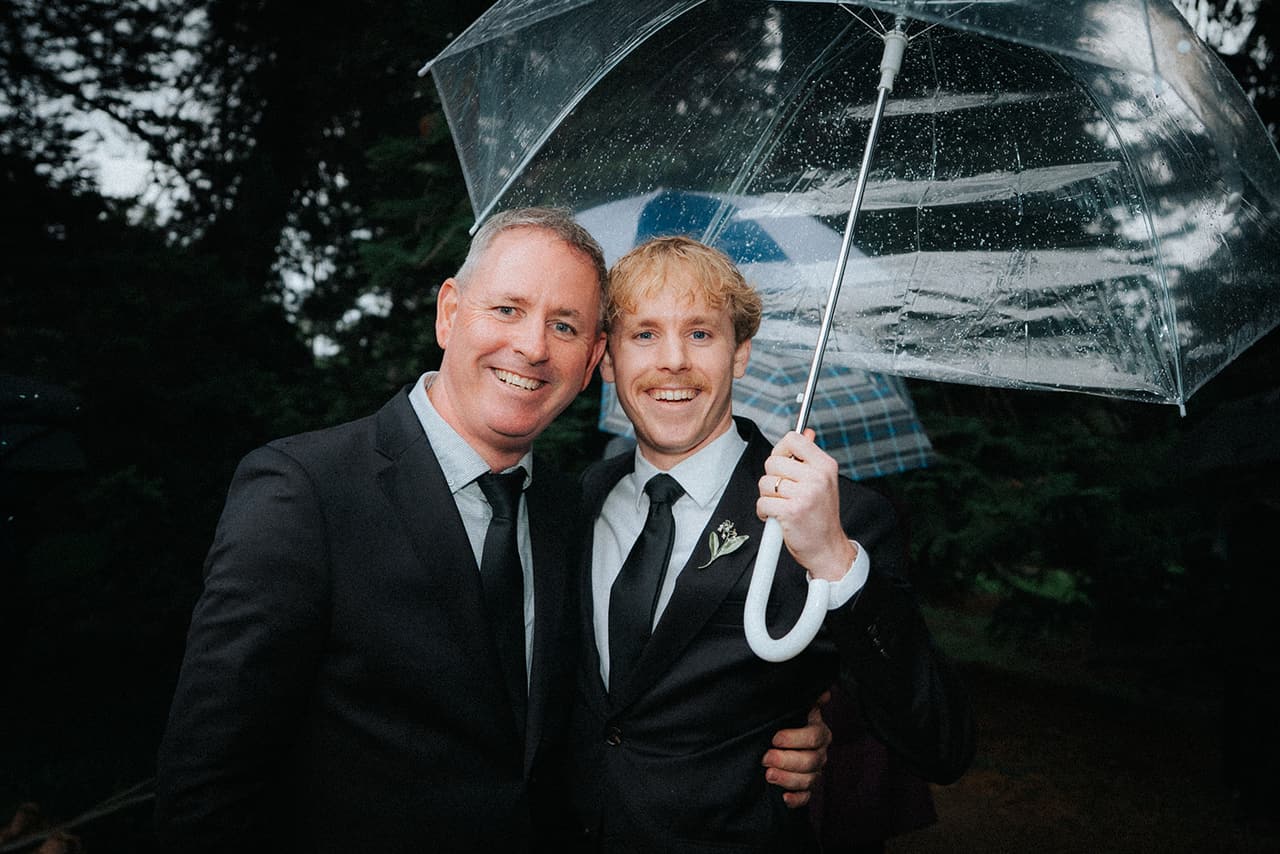 Eli holding a clear umbrella and grinning with a man in a dark suit who has an arm around him
