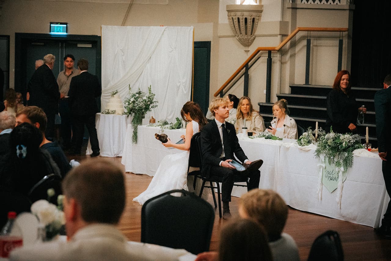 Charlotte and Eli sitting back to back with each other’s shoes in hand, playing the shoe game in front of the bridal table