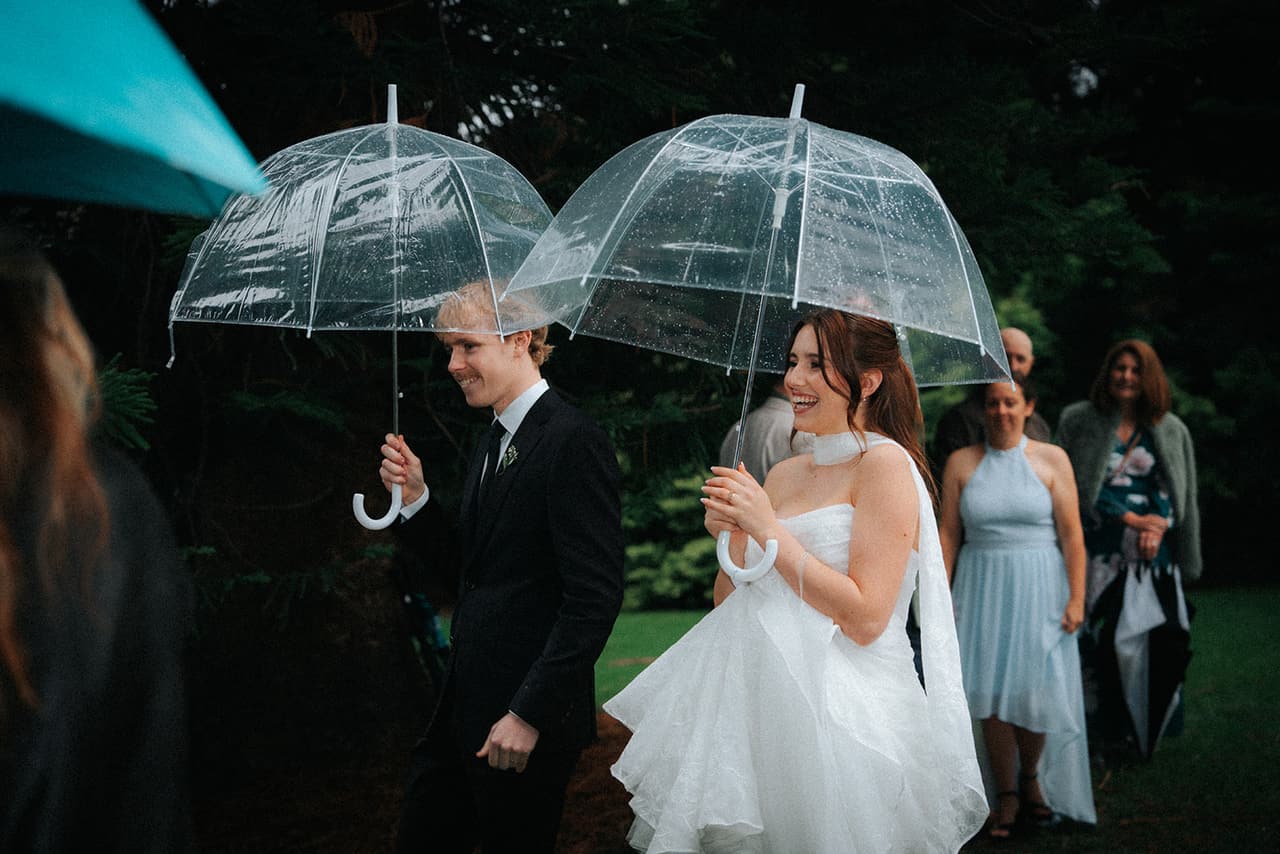 Eli and Charlotte each under a clear umbrella as they walk among their guests after the ceremony