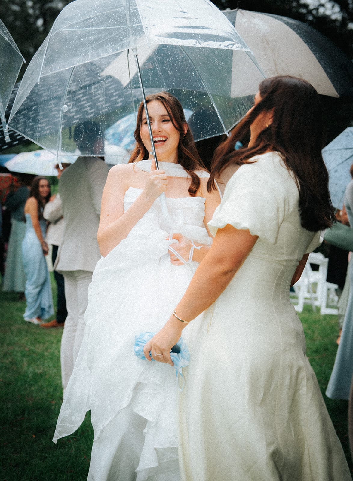 Charlotte laughing with a friend in a cream dress under clear umbrellas after the ceremony