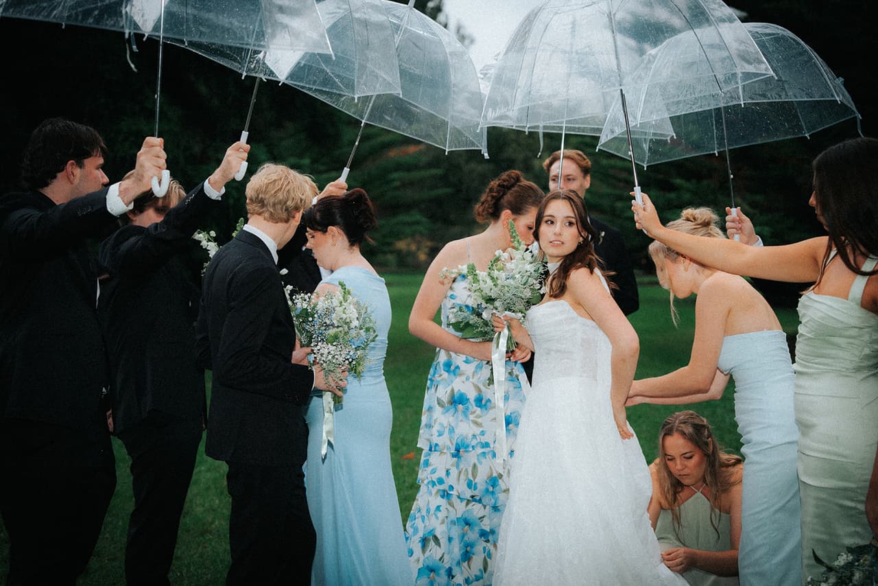 The bridal party holding clear umbrellas over Eli and Charlotte while a bridesmaid crouches to fix Charlotte’s dress