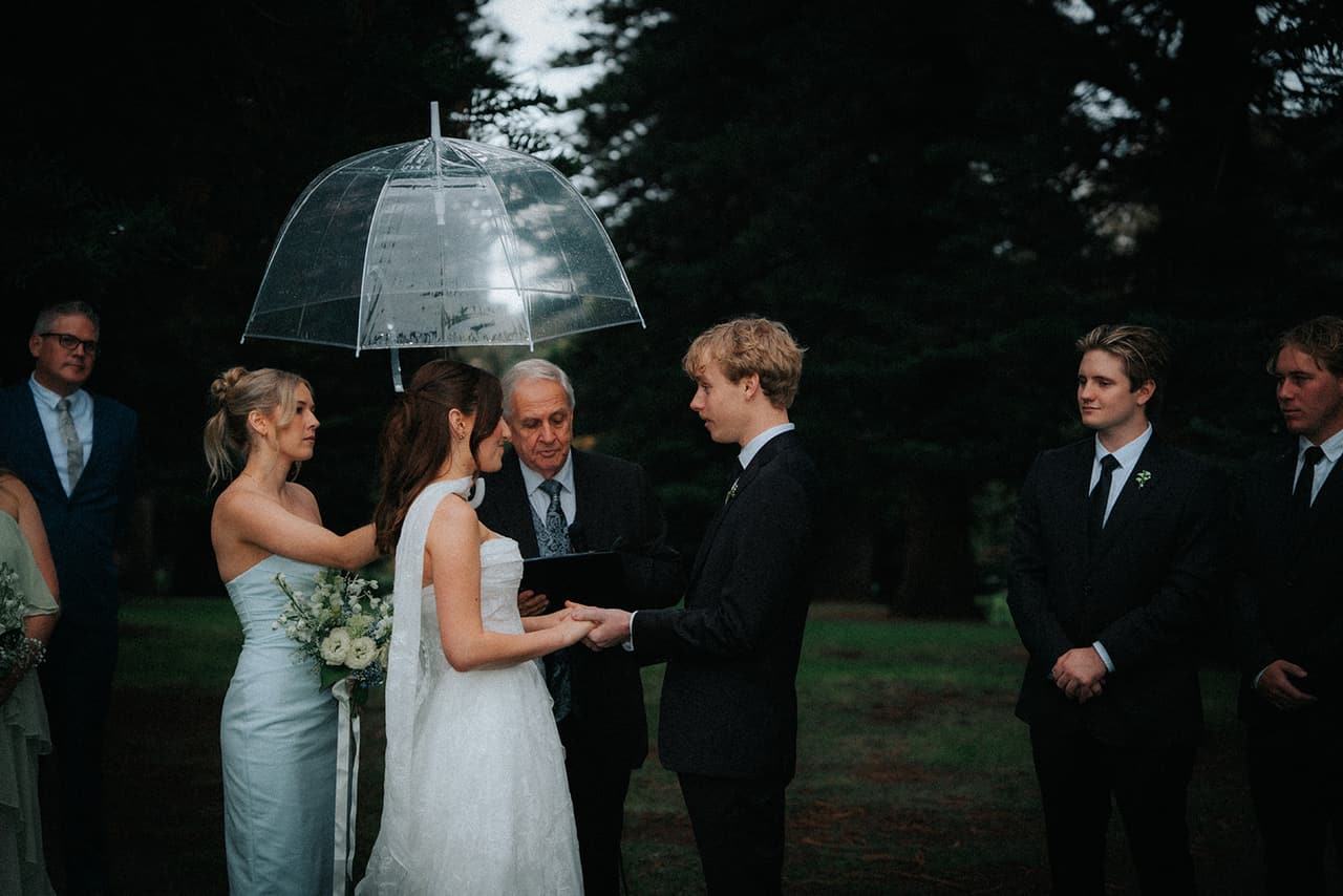 A bridesmaid holding a clear umbrella over Charlotte as she takes Eli’s hands in front of the celebrant