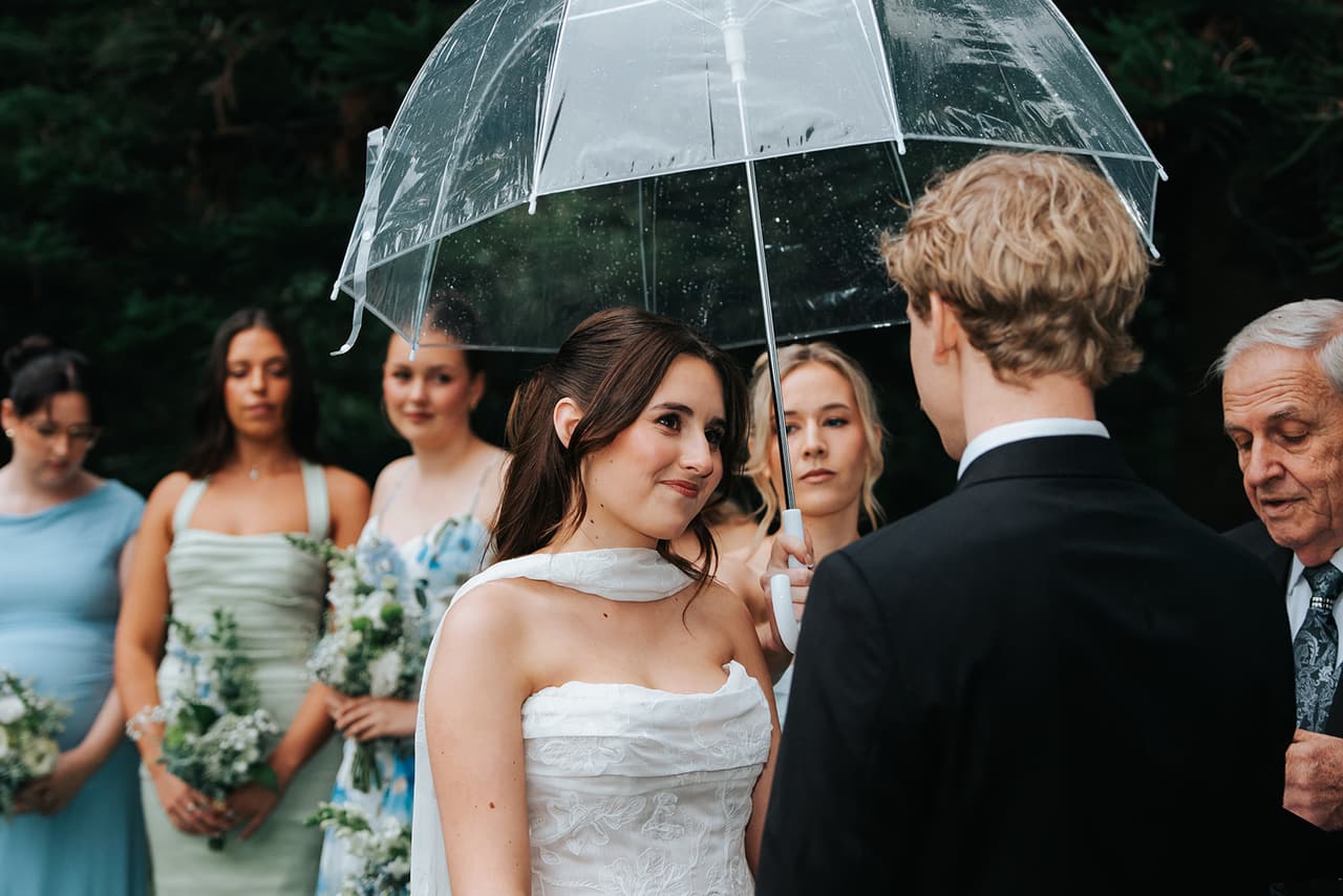 Charlotte smiling at Eli under a clear umbrella, her bridesmaids in blue, sage and floral dresses behind her