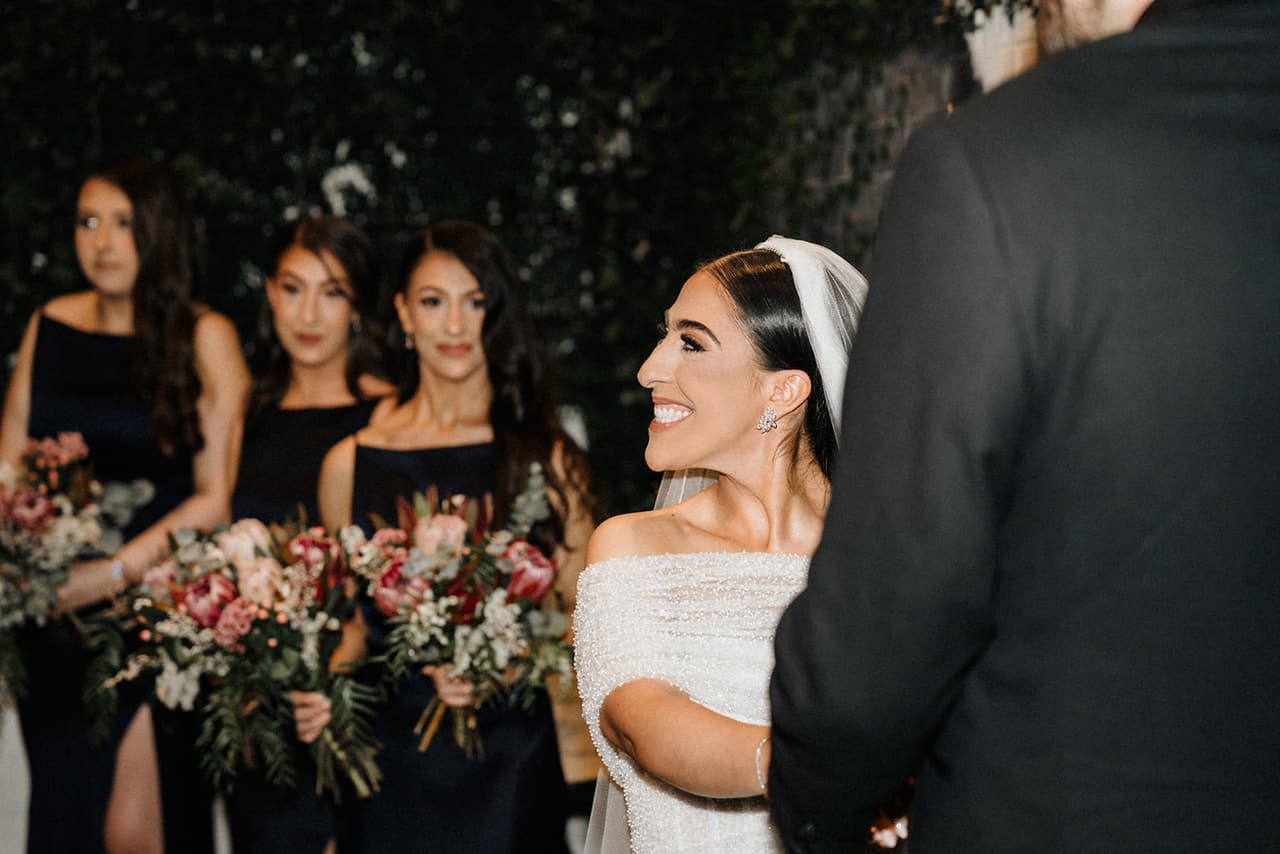 Tia beaming at George during the ceremony, her three bridesmaids in navy behind her with their bouquets