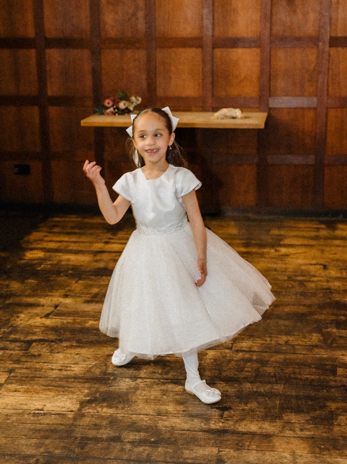 The flower girl twirling in her white dress on the floorboards of the timber-panelled room