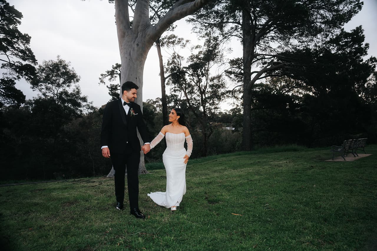 George and Tia walking hand in hand across a lawn beneath tall gums