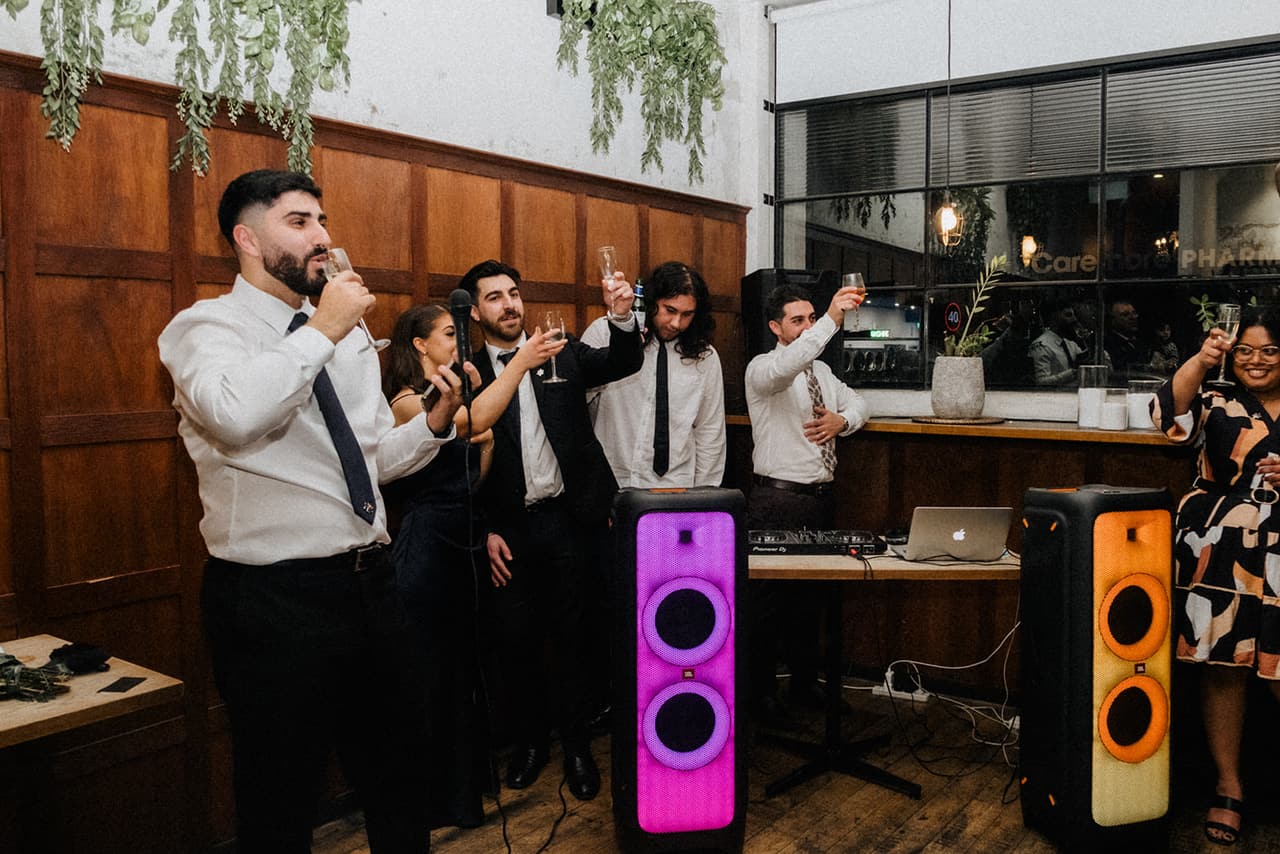 Groomsmen in white shirts raising their glasses in a toast beside the glowing party speakers