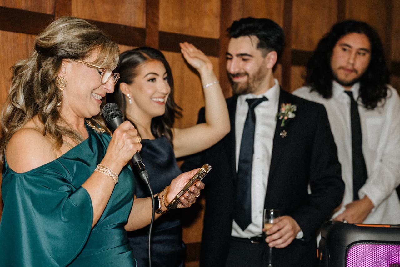 A woman in green giving a speech into a microphone as a bridesmaid and two young men laugh beside her