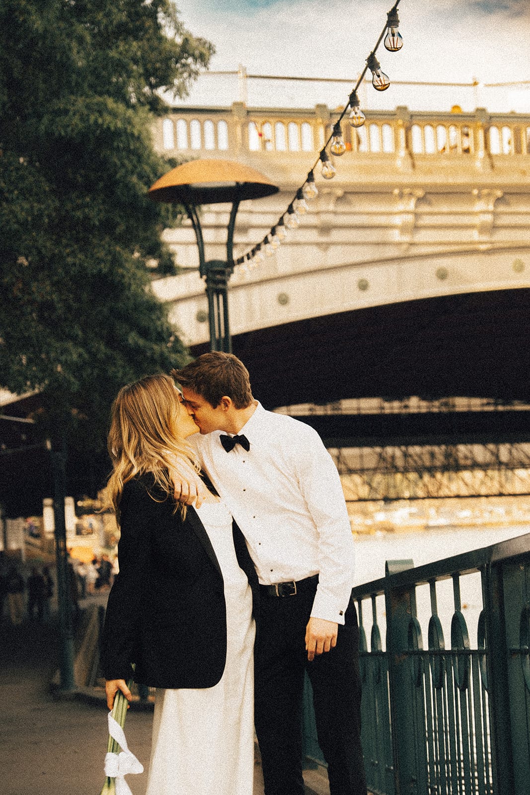 Hannah and Josh kissing on the riverside path beneath the balustrade of the bridge, festoon lights overhead