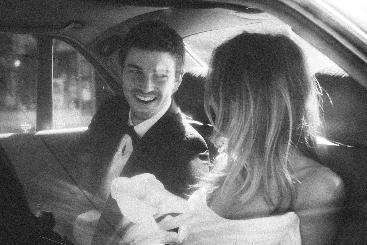 Black and white photo of Josh laughing as Hannah leans close in the back seat of the car