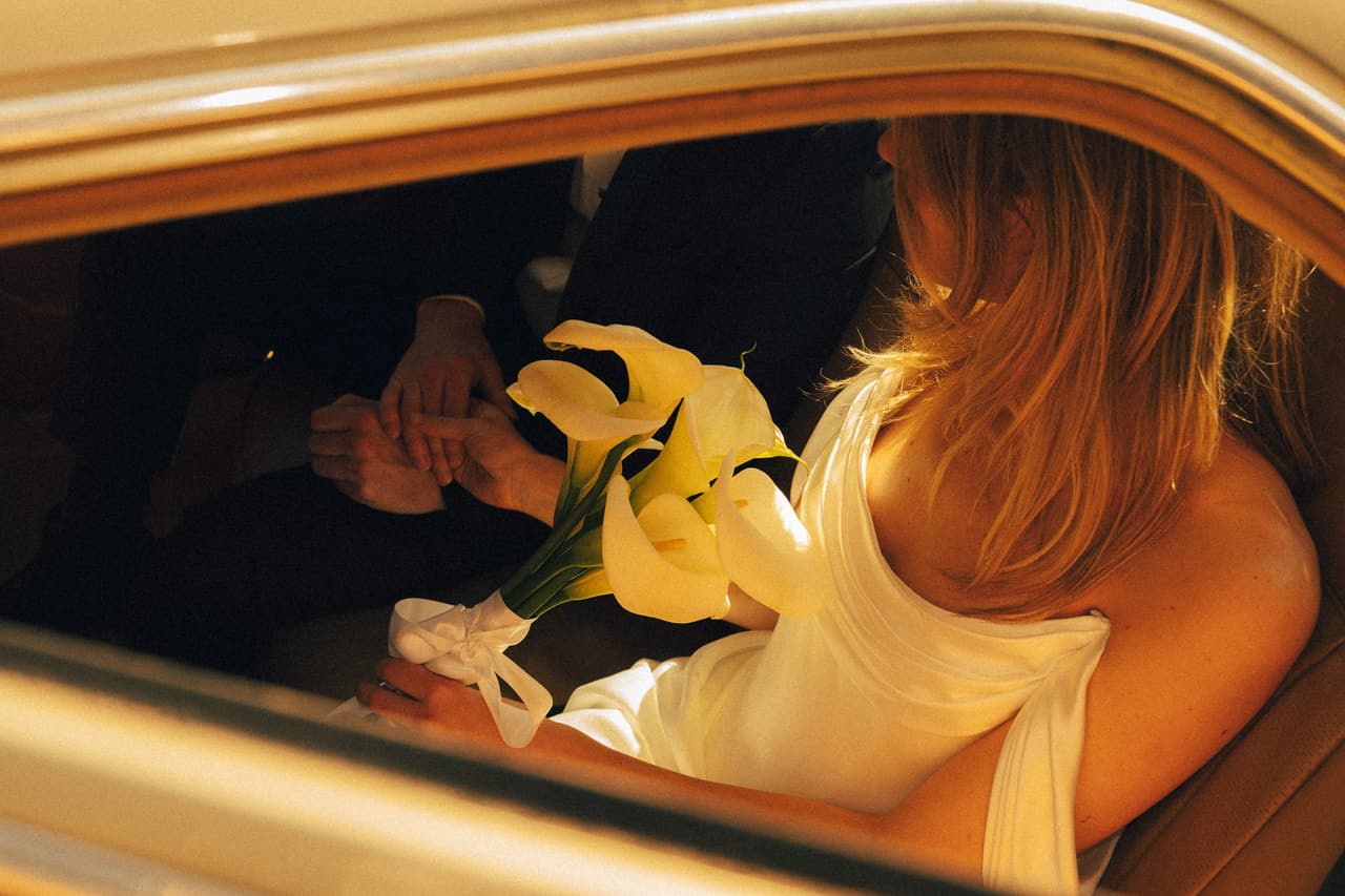 Hannah in the back of a vintage car holding yellow calla lilies, warm light on her gown