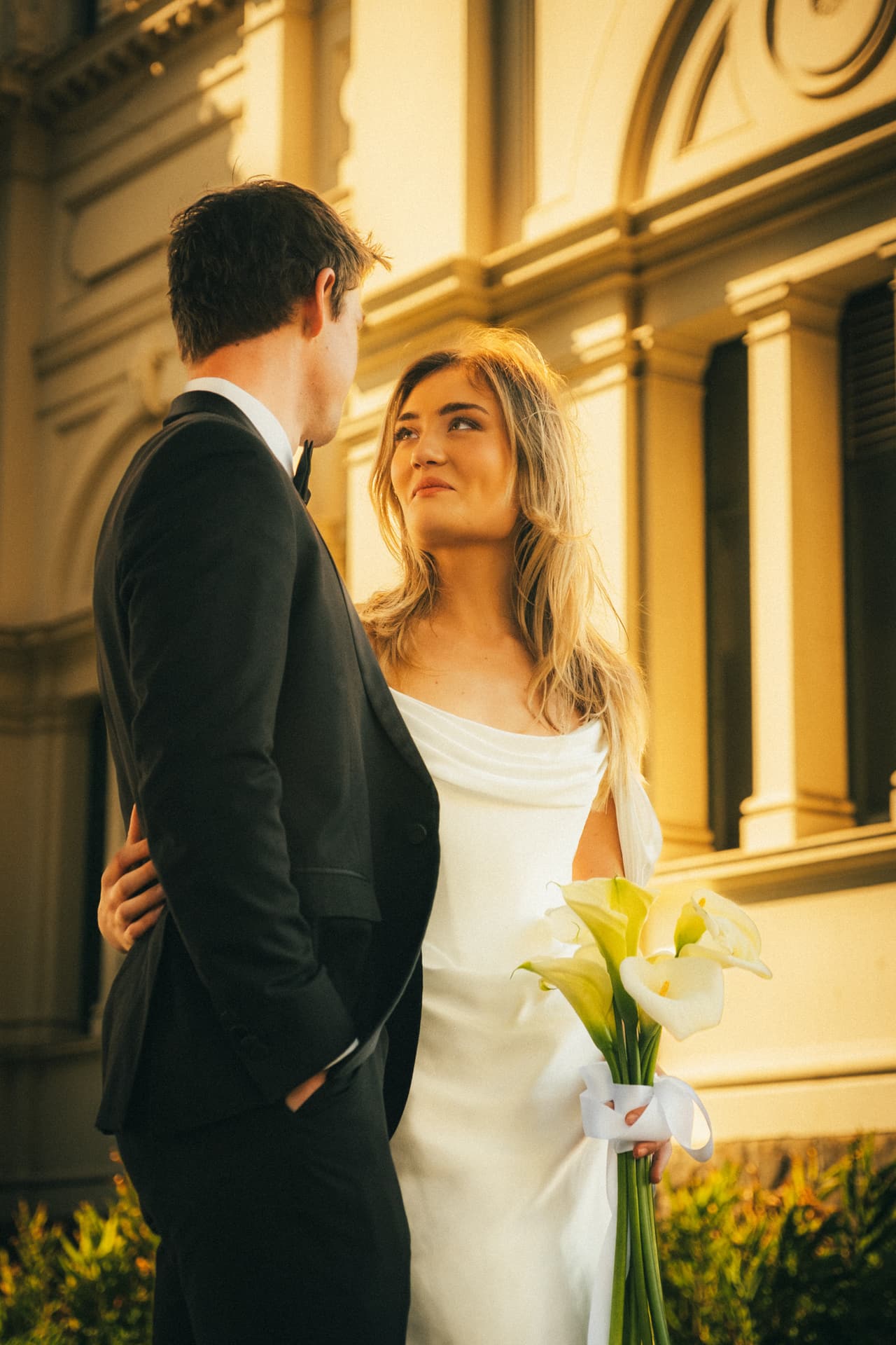 Hannah looking up at Josh in his dinner suit beside the Royal Exhibition Building in Carlton Gardens, calla lilies tied with white ribbon in her hand