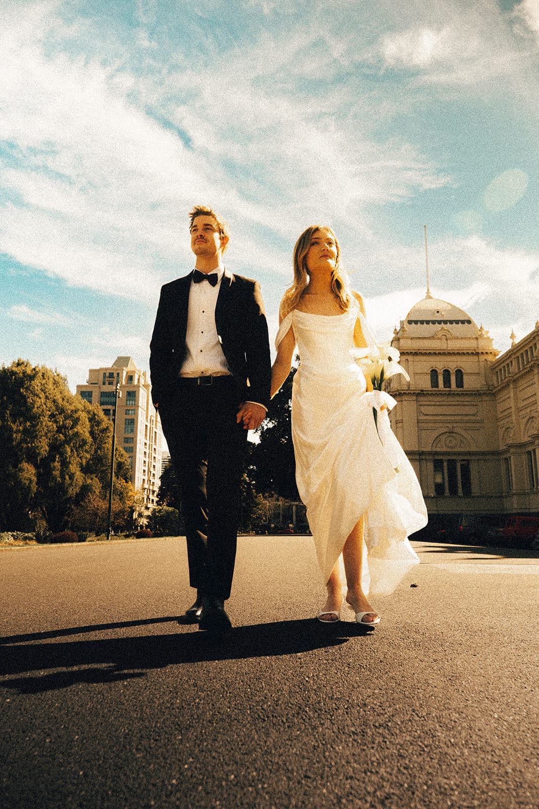 Hannah and Josh walking hand in hand on a path in Carlton Gardens, the Royal Exhibition Building behind them
