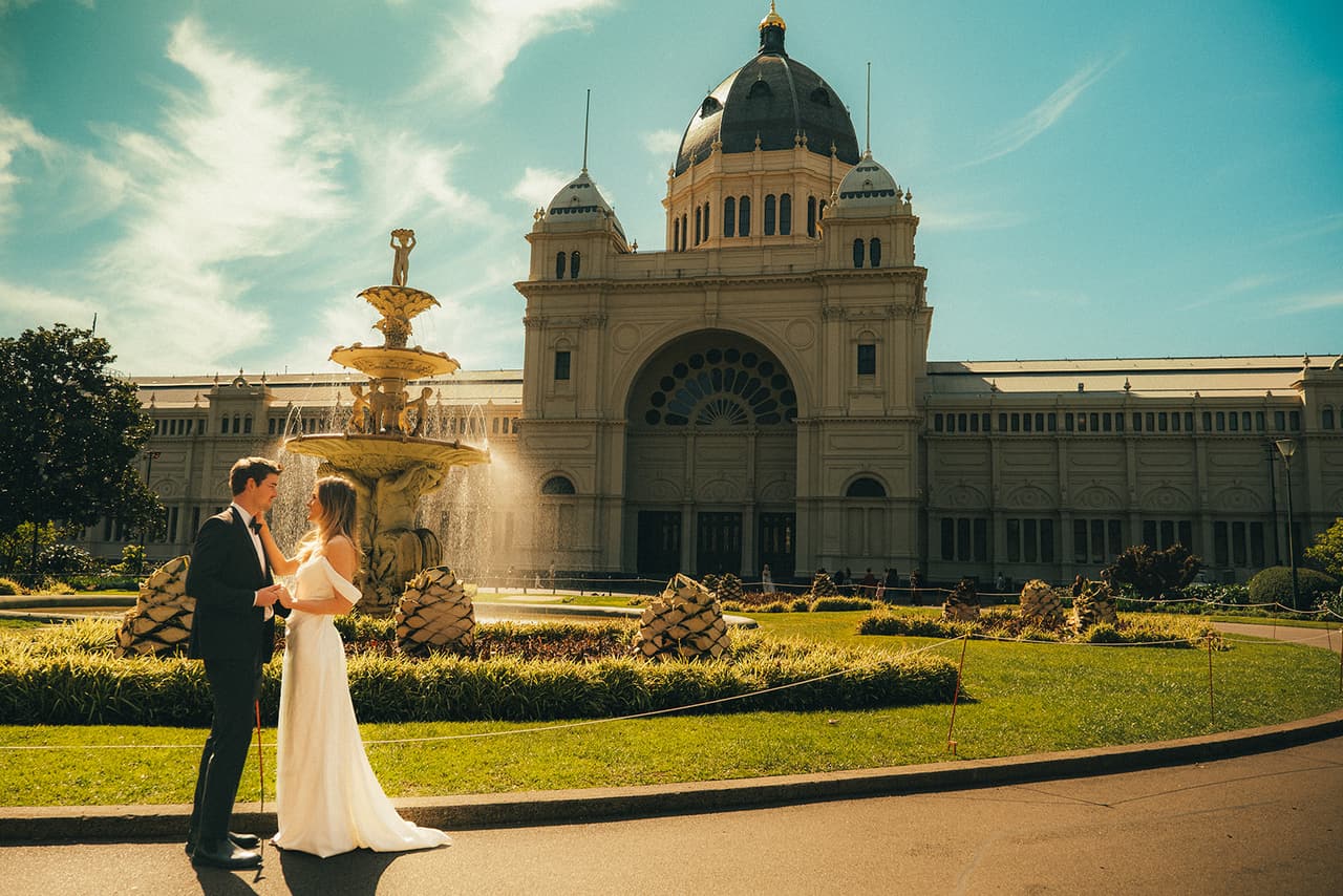 Hannah and Josh by the fountain in Carlton Gardens, the domed Royal Exhibition Building behind them under a blue sky