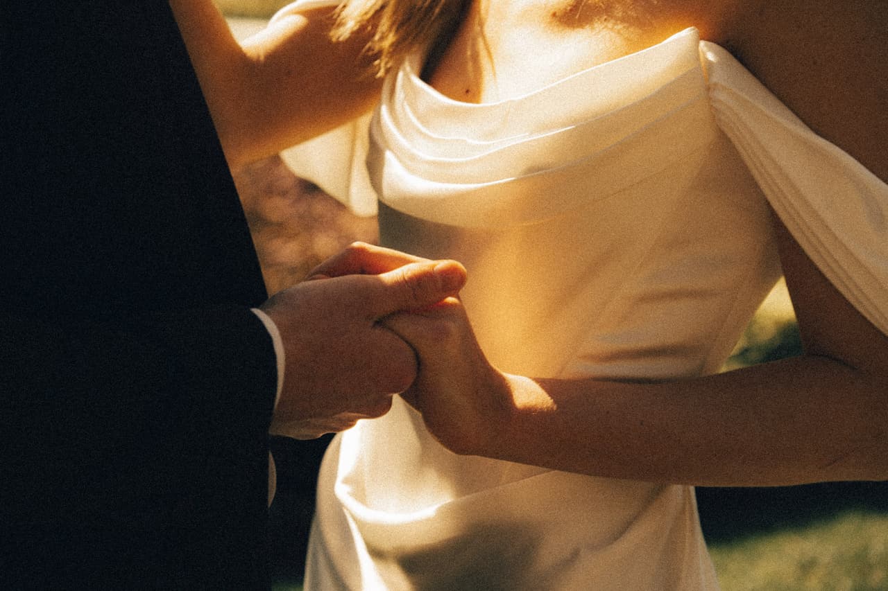 Close-up of Josh holding Hannah’s hand against her satin gown in late sun
