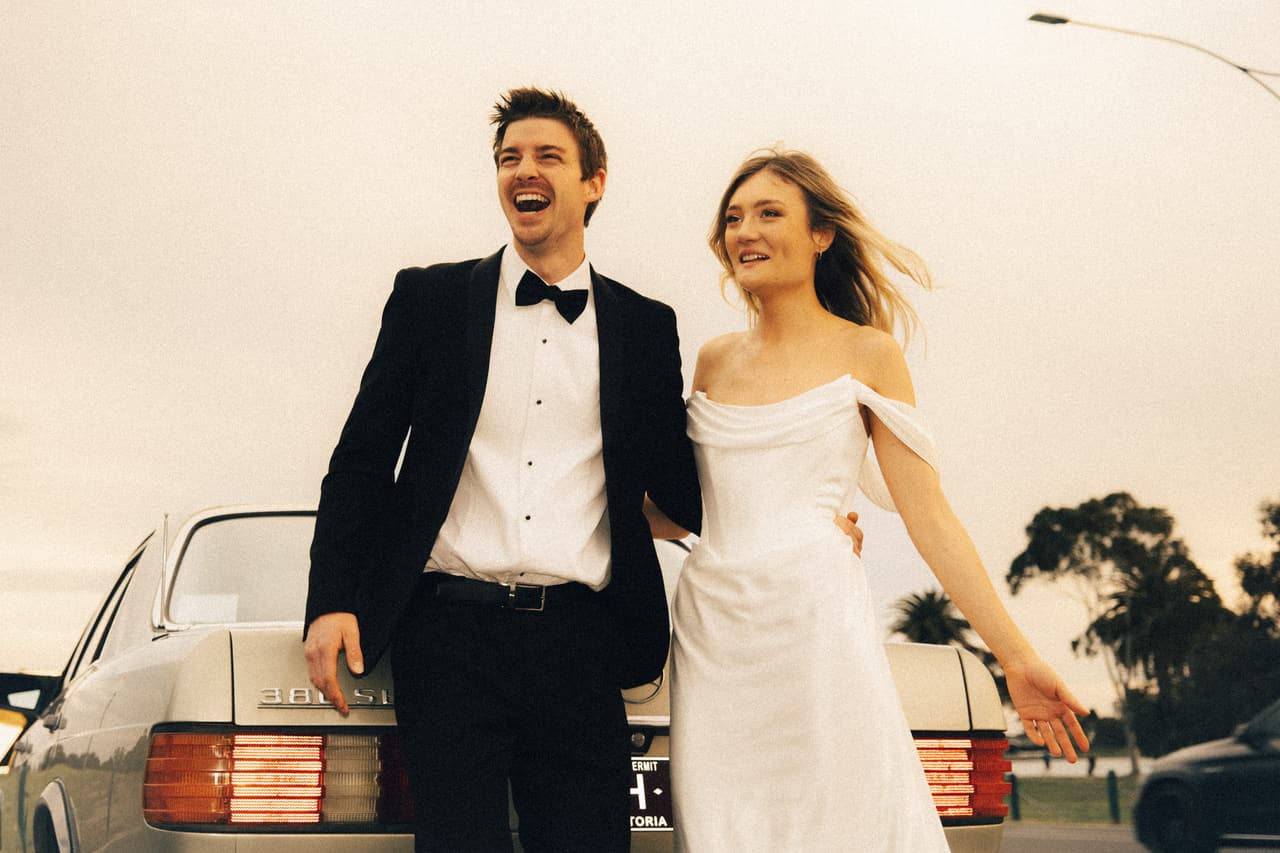 Josh laughing beside Hannah in front of a vintage Mercedes on a windy day