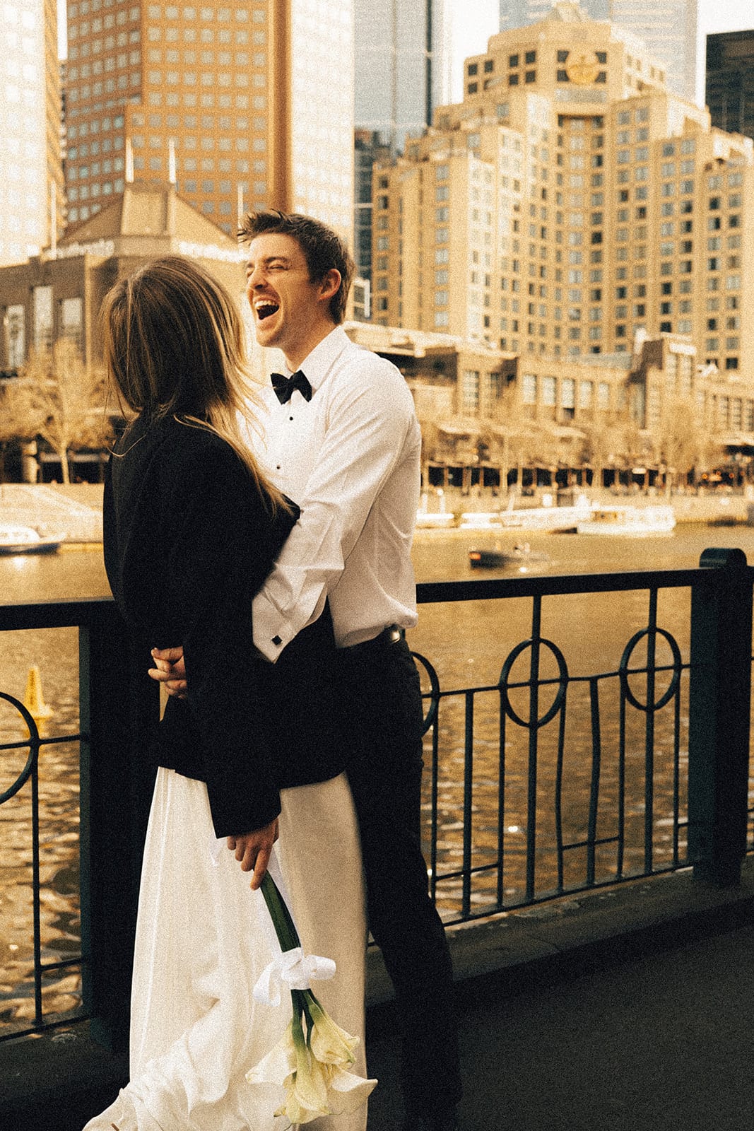 Josh laughing with Hannah, in his jacket, at the railing above the Yarra, city buildings across the river