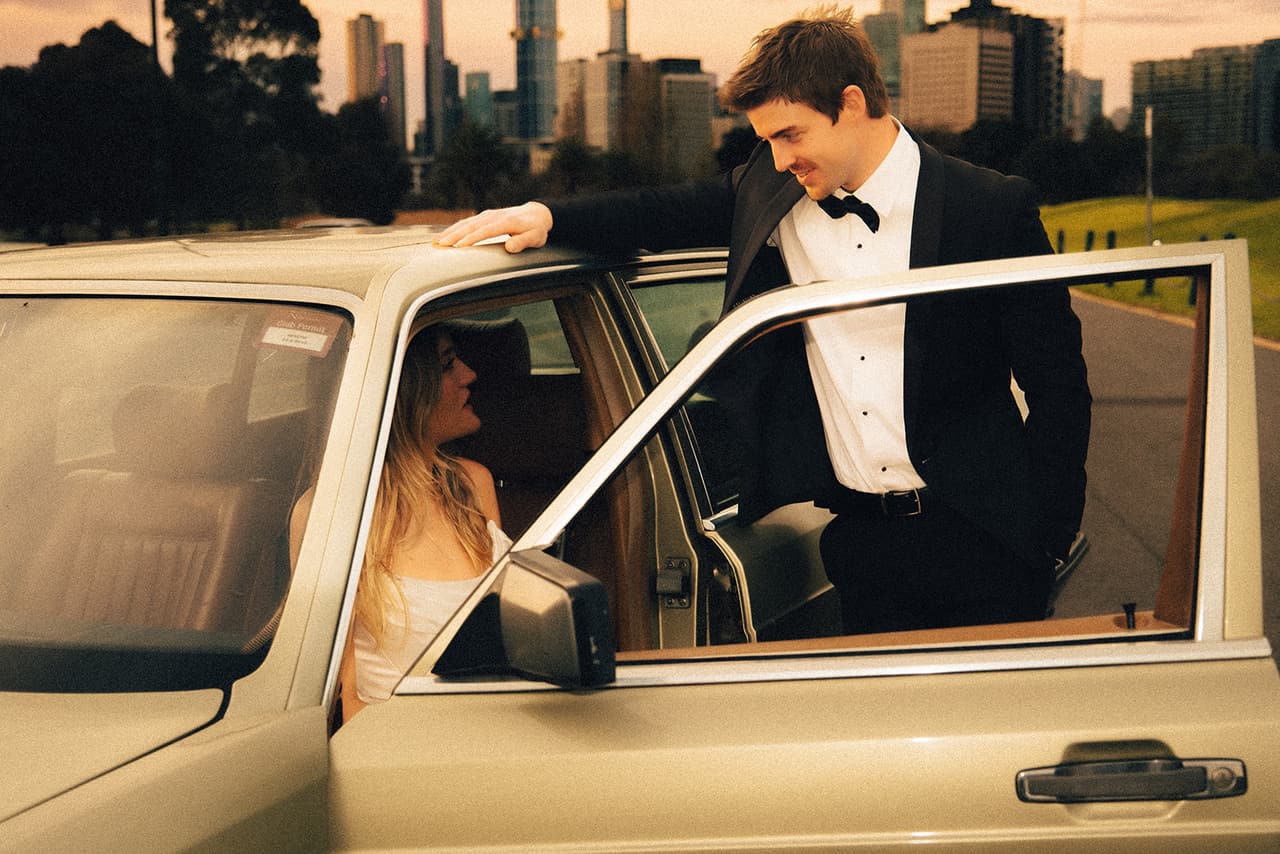 Josh leaning on the open door of the Mercedes with Hannah inside, the city skyline beyond the park