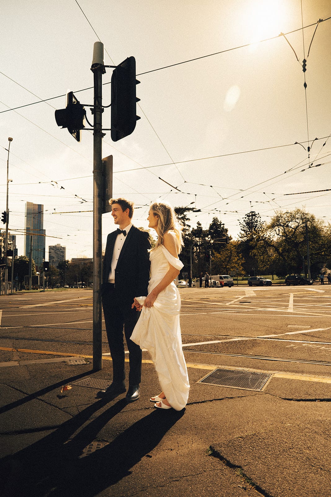 Hannah and Josh waiting by the traffic lights at a sunny intersection, tram wires overhead