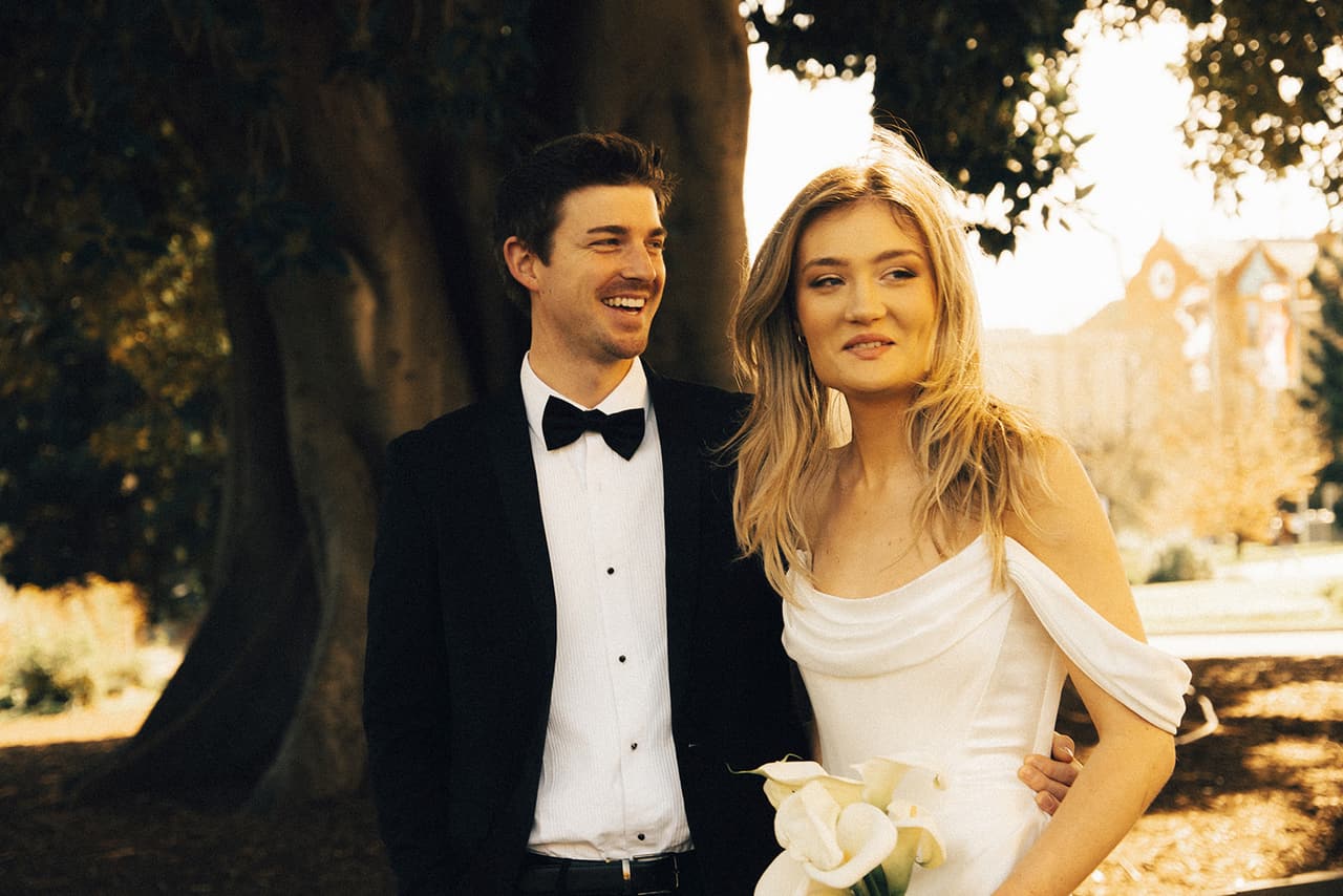 Josh laughing beside Hannah in the shade of a big tree, the sunlit gardens behind them
