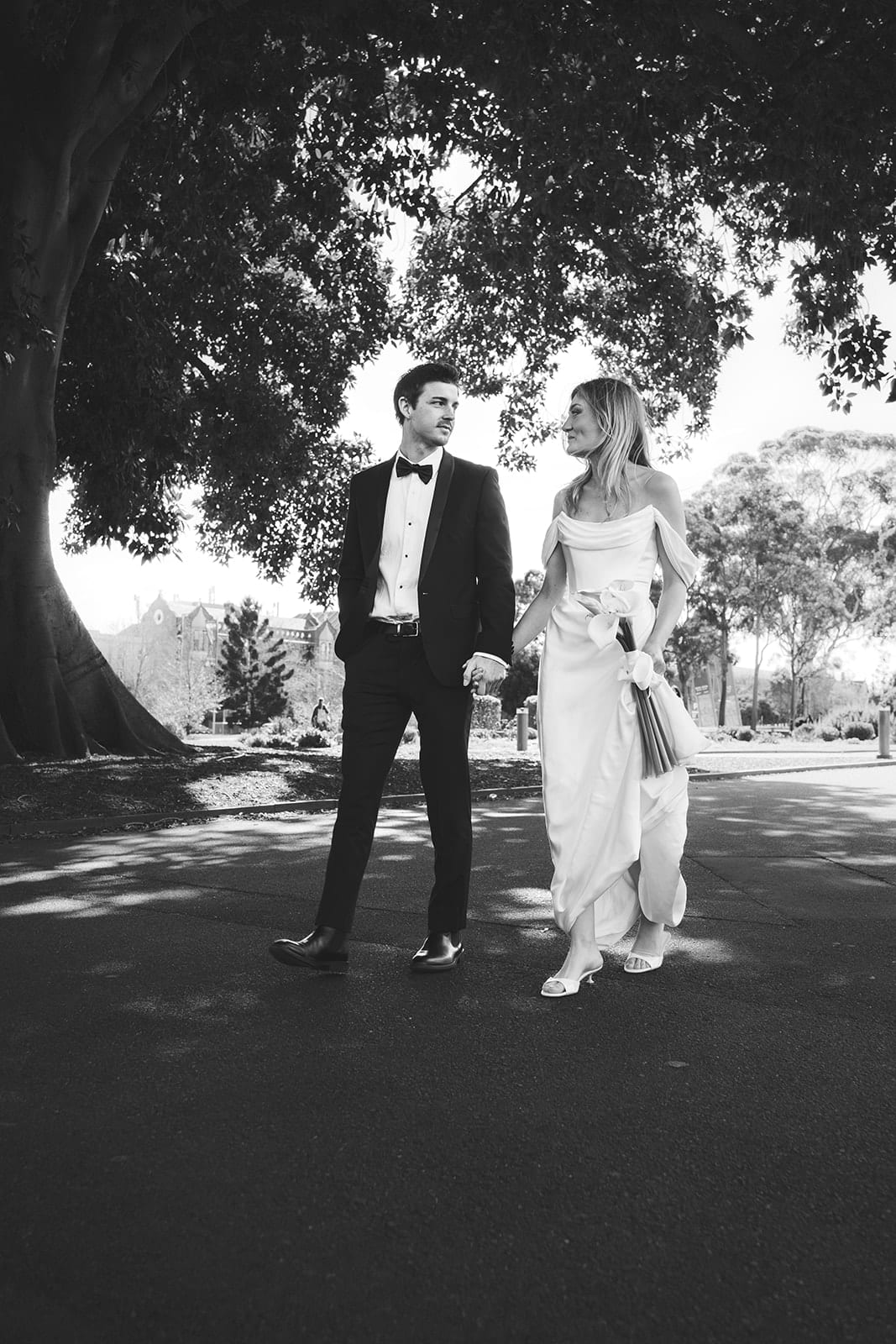 Black and white photo of Hannah and Josh walking towards the camera under the dark arch of a big tree in Carlton Gardens