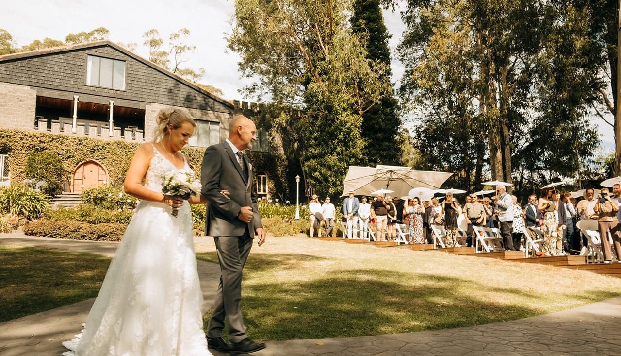 Jenna walking along the garden path to the ceremony arm in arm with an older man in a grey suit, the guests watching